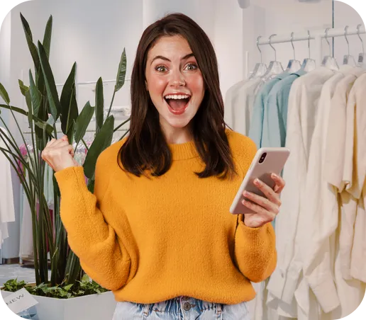 Vendedora de loja de roupas comemorando com o celular na mão, ao lado do estoque de peças, usando o CRM com WhatsApp do ConnectStore
